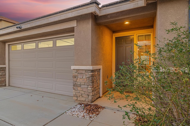 exterior entry at dusk featuring an attached garage, stone siding, and stucco siding