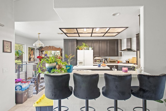 kitchen with white refrigerator with ice dispenser, wall chimney range hood, light tile patterned floors, a kitchen bar, and kitchen peninsula