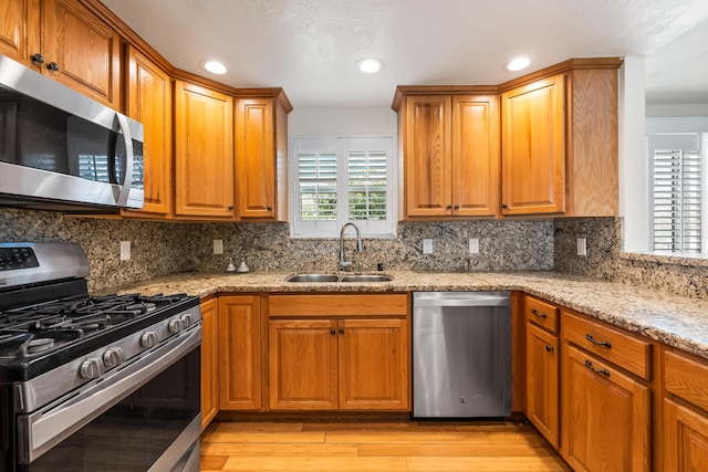 kitchen with appliances with stainless steel finishes, light wood-style floors, brown cabinetry, a sink, and light stone countertops