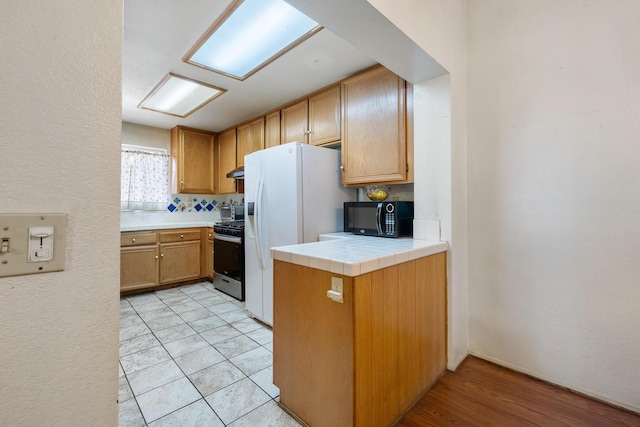 kitchen featuring stainless steel gas range oven, light tile patterned floors, tile counters, kitchen peninsula, and white refrigerator with ice dispenser