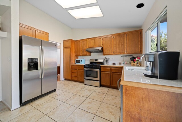 kitchen with brown cabinets, light tile patterned floors, stainless steel appliances, a sink, and under cabinet range hood