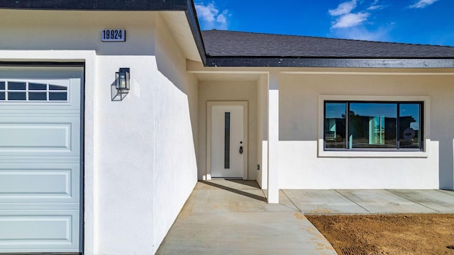 entrance to property with stucco siding, a garage, and roof with shingles