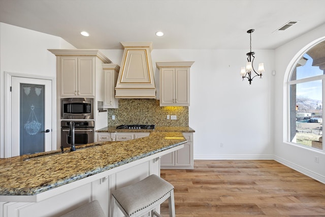 kitchen featuring dark stone countertops, cream cabinetry, decorative backsplash, custom range hood, and appliances with stainless steel finishes