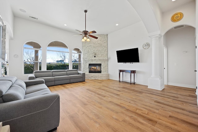 living room featuring light wood-type flooring, a towering ceiling, ornate columns, ceiling fan, and a stone fireplace