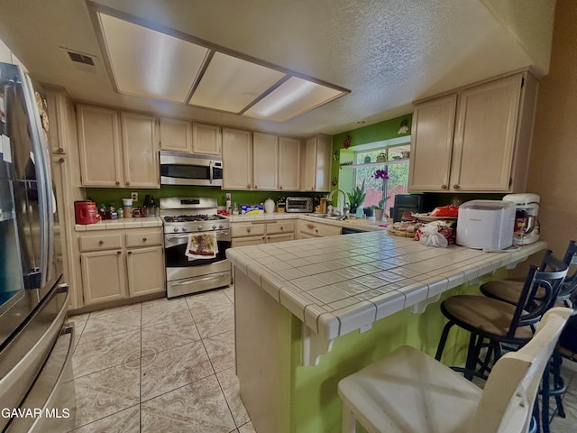 1816 Orange St, Rosamond, CA 93560 - kitchen with a peninsula, appliances with stainless steel finishes, tile counters, a breakfast bar area, and light brown cabinets