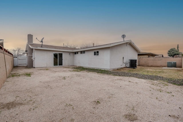 back of house at dusk featuring central air condition unit, a fenced backyard, a chimney, and stucco siding