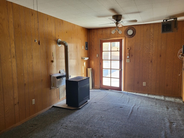 interior space featuring dark colored carpet, a wood stove, wooden walls, and ceiling fan