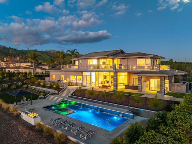 back house at dusk featuring a fire pit, a patio area, a balcony, and a swimming pool with hot tub