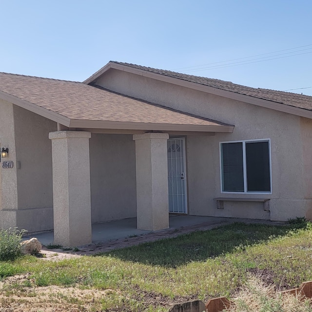 doorway to property with a yard, stucco siding, and roof with shingles