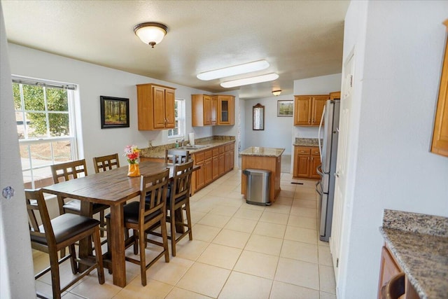 12332 E Ave X, Pearblossom, CA 93553 - kitchen featuring freestanding refrigerator, brown cabinetry, light tile patterned floors, light stone counters, and glass insert cabinets