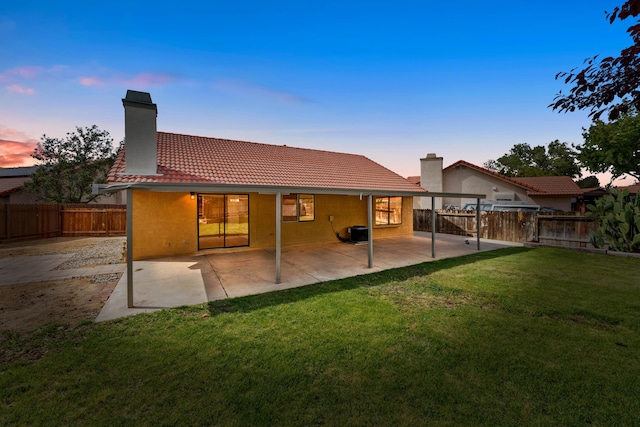 back of property at dusk with a tiled roof, a chimney, a fenced backyard, a yard, and stucco siding
