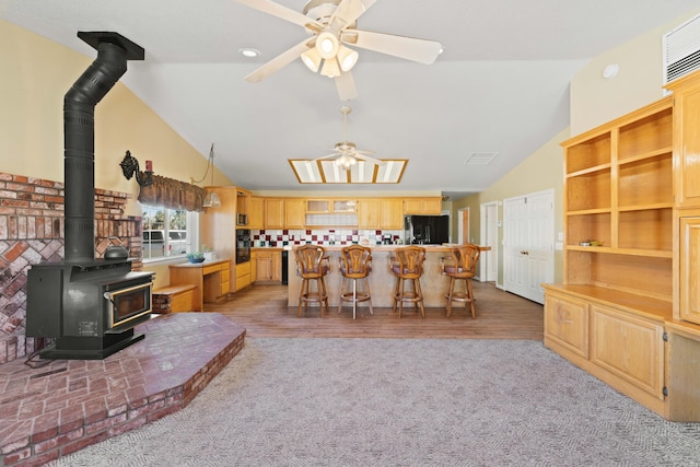 kitchen featuring ceiling fan, vaulted ceiling, light brown cabinetry, black appliances, and a wood stove