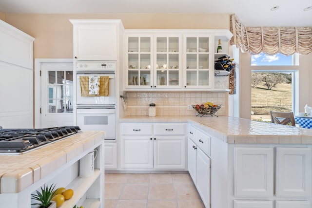 kitchen featuring white double oven, tile counters, stainless steel gas cooktop, white cabinets, and decorative backsplash