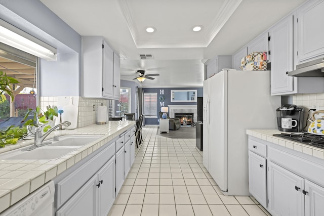 kitchen with a raised ceiling, sink, a brick fireplace, tile counters, and white cabinetry