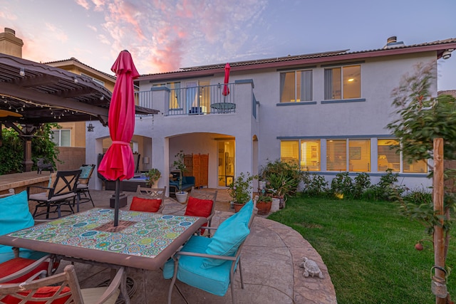 back house at dusk featuring a pergola, a patio area, a balcony, and a lawn