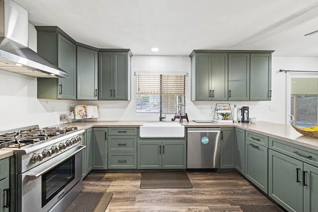 kitchen with stainless steel appliances, green cabinetry, a sink, and wall chimney exhaust hood