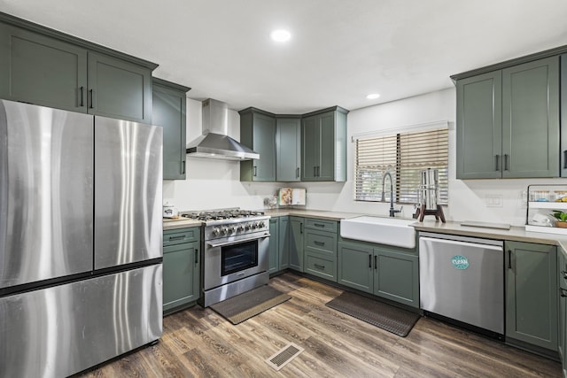 kitchen featuring wall chimney range hood, light countertops, a sink, green cabinetry, and stainless steel appliances