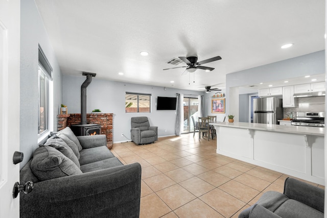 living room featuring light tile patterned floors, visible vents, baseboards, ceiling fan, and a wood stove