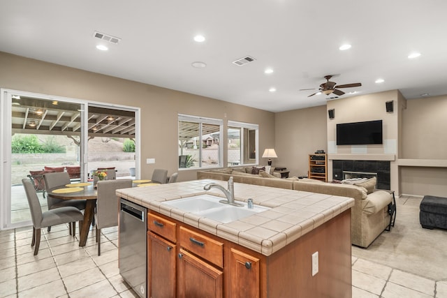 kitchen featuring tile countertops, a kitchen island with sink, a tile fireplace, sink, and stainless steel dishwasher