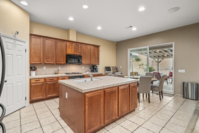 kitchen with tile countertops, backsplash, a kitchen island with sink, sink, and light tile patterned floors