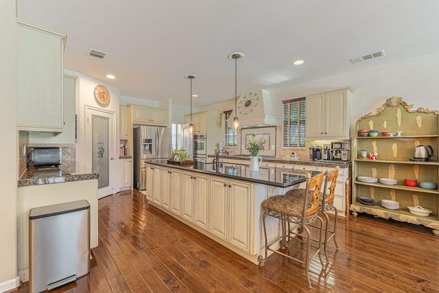 kitchen featuring custom exhaust hood, a kitchen island with sink, appliances with stainless steel finishes, cream cabinetry, and decorative light fixtures