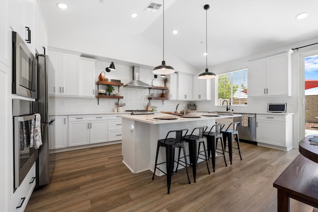 kitchen with wall chimney range hood, appliances with stainless steel finishes, a breakfast bar, open shelves, and wood finished floors