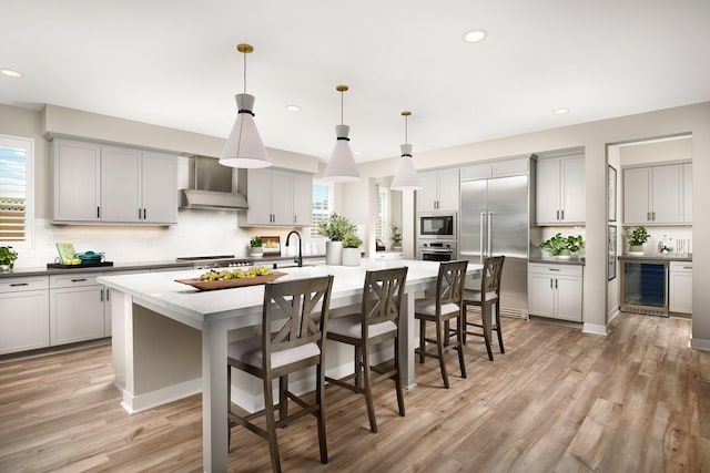 kitchen featuring a kitchen island with sink, hanging light fixtures, built in appliances, wall chimney exhaust hood, and gray cabinets