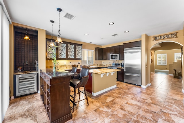 kitchen with beverage cooler, arched walkways, stainless steel appliances, dark brown cabinetry, and a center island