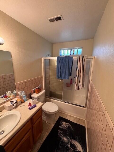 full bathroom with vanity, tile walls, toilet, visible vents, and enclosed tub / shower combo