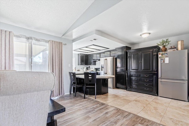 kitchen with dark cabinetry, a breakfast bar area, freestanding refrigerator, light countertops, and a textured ceiling