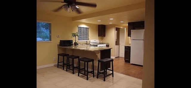 kitchen with a kitchen bar, kitchen peninsula, ceiling fan, white appliances, and light stone counters