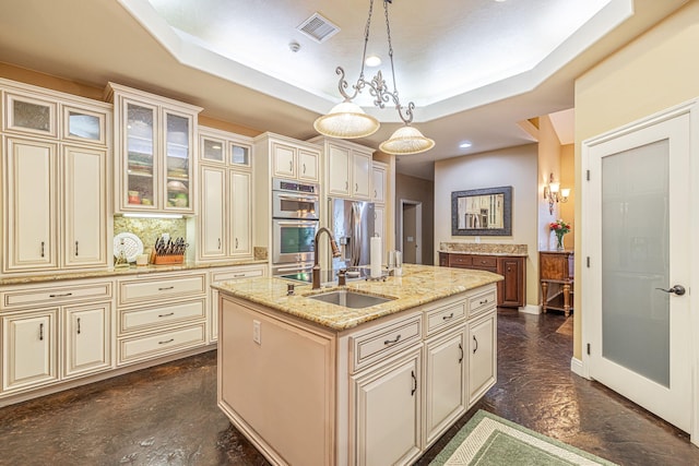 kitchen with a raised ceiling, light stone counters, pendant lighting, cream cabinetry, and a center island with sink