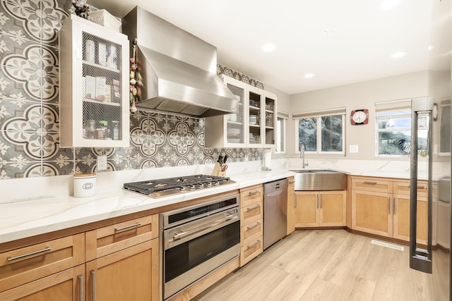 kitchen with light brown cabinets, wall chimney range hood, light stone counters, stainless steel appliances, and a sink