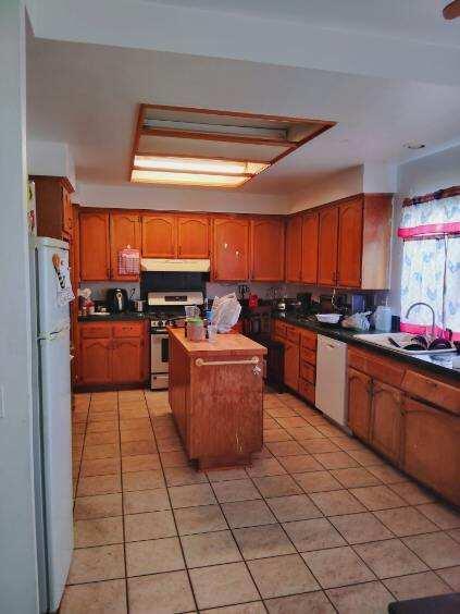 kitchen with brown cabinetry, light tile patterned flooring, a kitchen island, white appliances, and under cabinet range hood