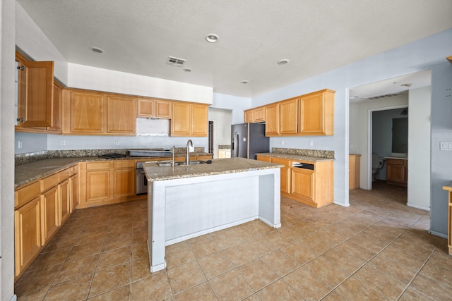 kitchen featuring a kitchen island with sink, light stone counters, stainless steel fridge, and visible vents