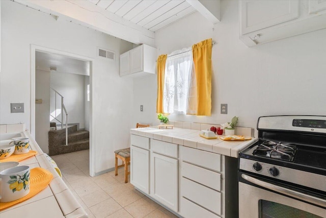 kitchen featuring stainless steel gas range oven, visible vents, white cabinets, tile countertops, and beam ceiling