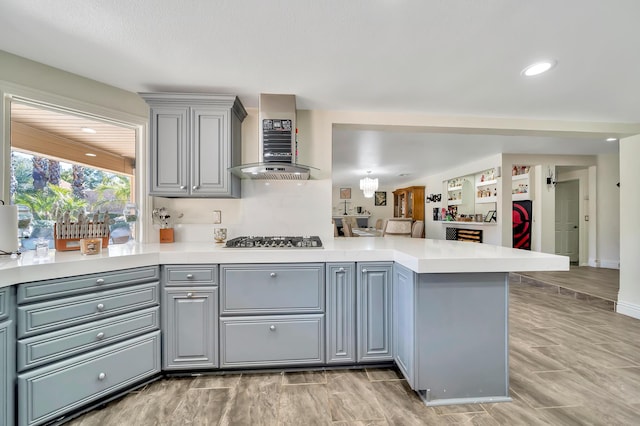 kitchen featuring light countertops, stainless steel gas stovetop, gray cabinetry, a peninsula, and wall chimney exhaust hood