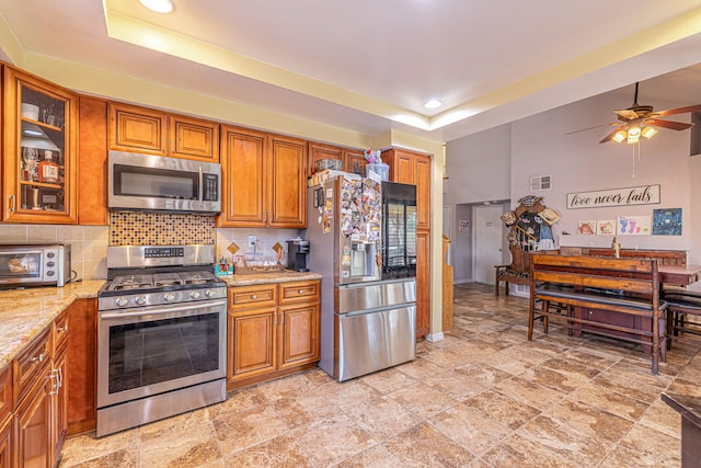 kitchen featuring a raised ceiling, ceiling fan, decorative backsplash, appliances with stainless steel finishes, and light stone counters