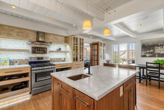 kitchen featuring stainless steel gas range, sink, wall chimney range hood, beam ceiling, and an island with sink