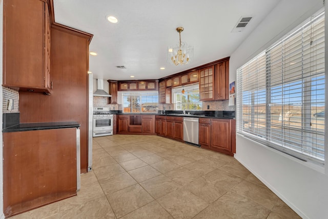 kitchen featuring hanging light fixtures, appliances with stainless steel finishes, wall chimney exhaust hood, dark countertops, and glass insert cabinets