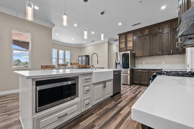 kitchen with appliances with stainless steel finishes, ornamental molding, dark brown cabinetry, a kitchen island with sink, and sink