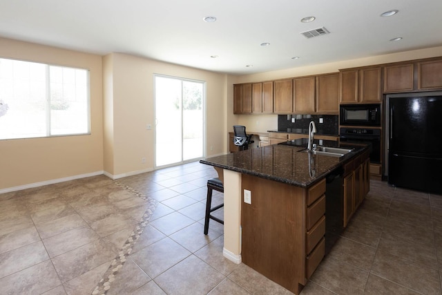 kitchen featuring sink, dark stone countertops, an island with sink, light tile patterned floors, and black appliances