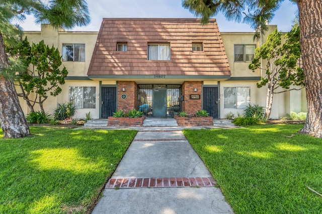 view of property featuring brick siding, mansard roof, a front yard, and stucco siding