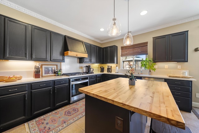 kitchen with custom range hood, appliances with stainless steel finishes, hanging light fixtures, wooden counters, and light tile patterned flooring