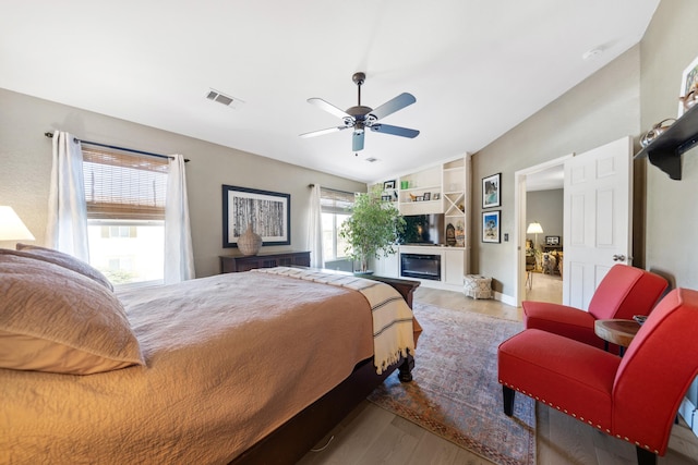 bedroom featuring visible vents, a ceiling fan, a glass covered fireplace, wood finished floors, and vaulted ceiling