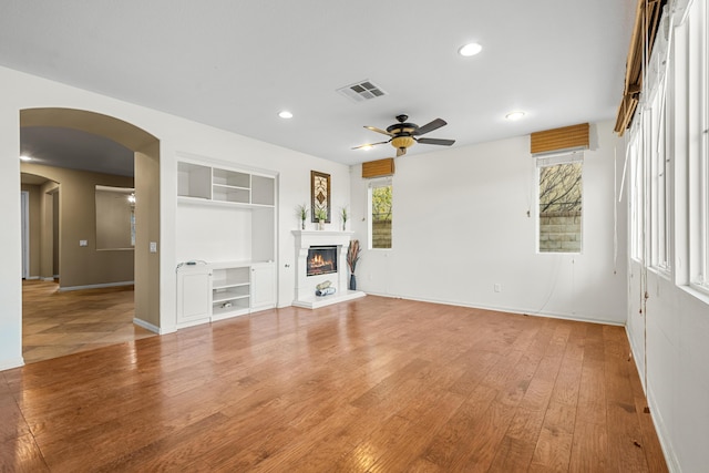 unfurnished living room featuring arched walkways, visible vents, light wood-style flooring, a ceiling fan, and a glass covered fireplace