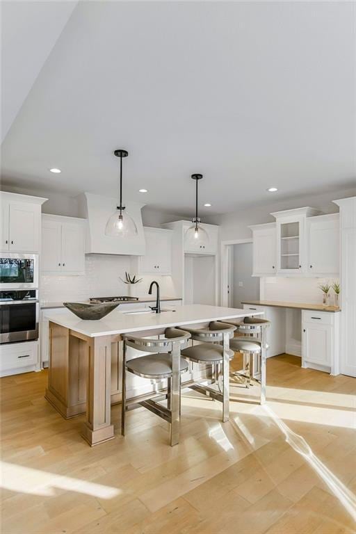 kitchen with light wood-type flooring, stainless steel appliances, a center island with sink, white cabinets, and hanging light fixtures