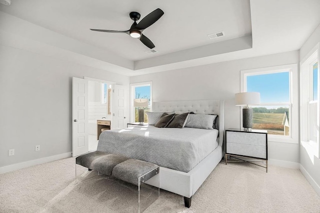 carpeted bedroom featuring a tray ceiling, multiple windows, ceiling fan, and ensuite bathroom