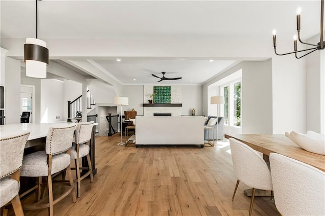 dining room with ceiling fan with notable chandelier, crown molding, light wood-type flooring, and a stone fireplace