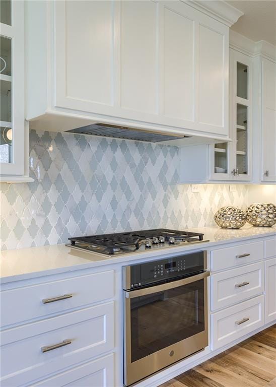 kitchen with appliances with stainless steel finishes, light wood-type flooring, tasteful backsplash, exhaust hood, and white cabinetry